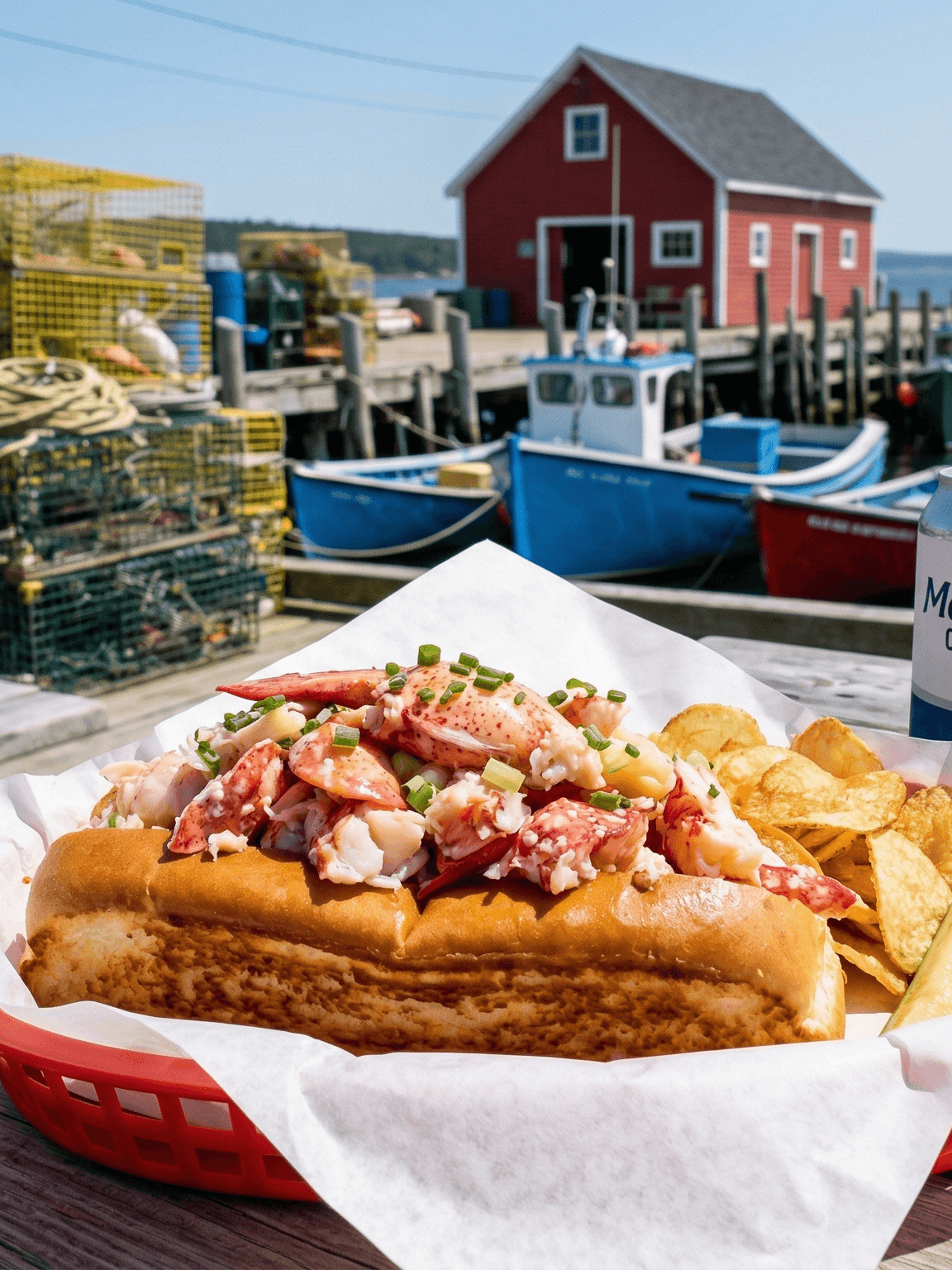 A fresh lobster roll with chips served at a wooden dock beside fishing boats and lobster traps in Nova Scotia, Canada